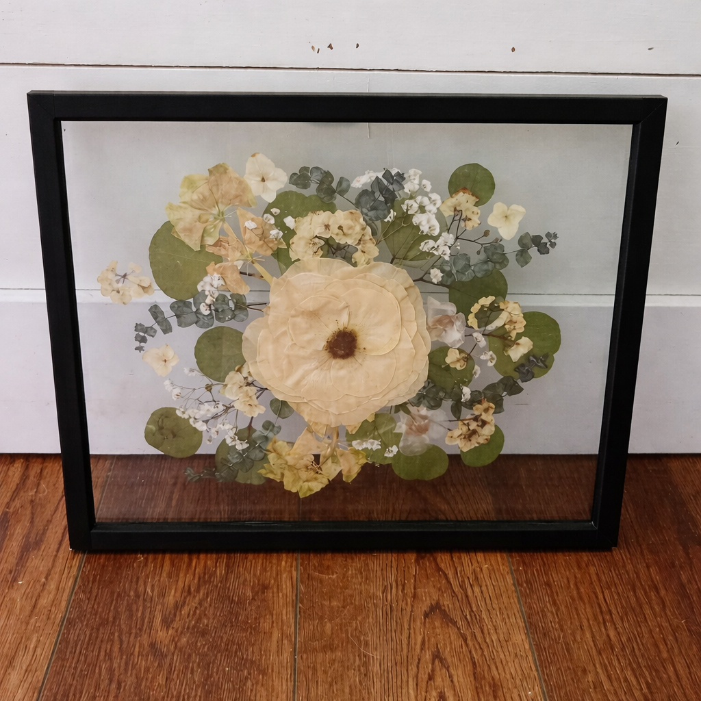 Framed pressed flowers on a wooden floor in a black frame with a white wall background