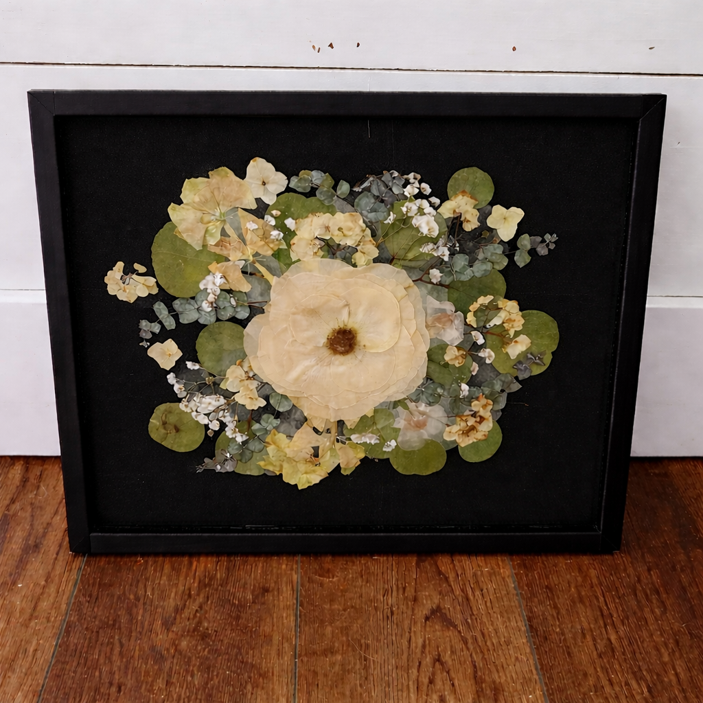 Framed pressed flowers in a black frame on a black mat on a wooden floor with a white wall background