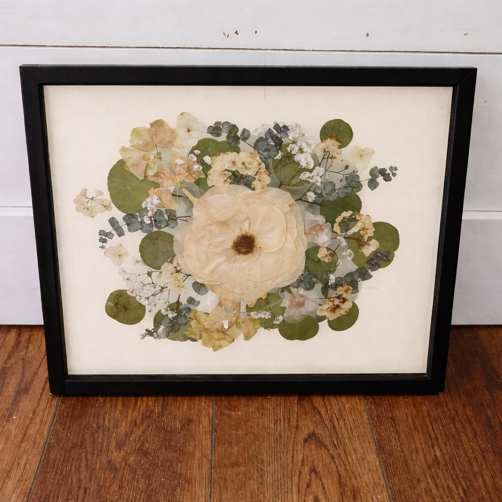 Framed pressed flower arrangement in a black frame and an ivory mat on a wooden floor with a white wall background