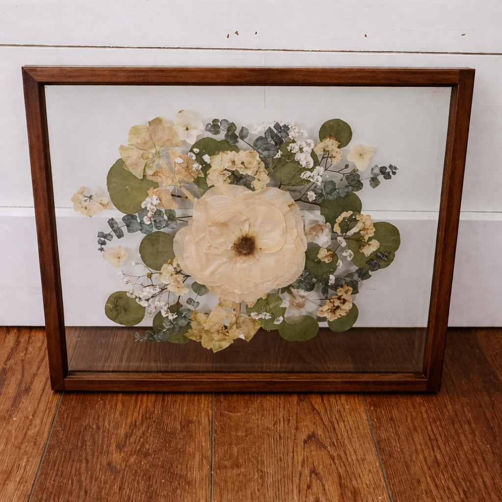 Framed pressed flowers in a double floating glass frame in a dark walnut stain on a wooden floor with a white wall background