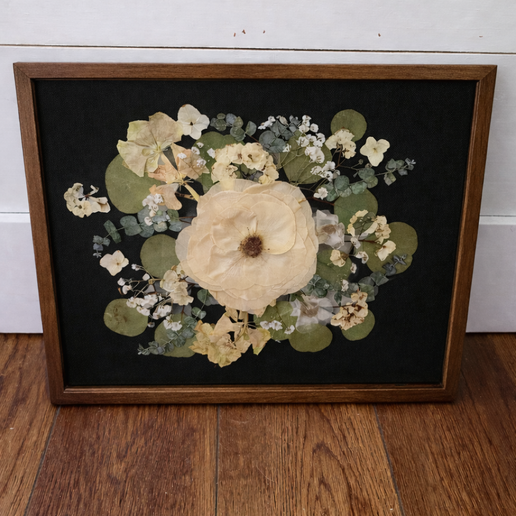 Framed pressed flowers on a black background in a dark walnut frame on a wooden floor with a white wall background
