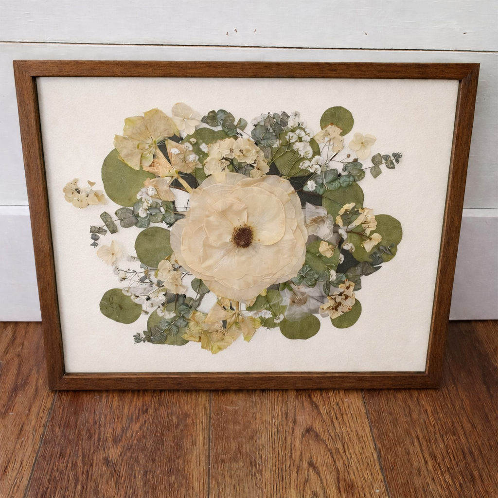 Framed pressed flower arrangement in a dark walnut frame on an ivory mat on a wooden floor with a white wall background