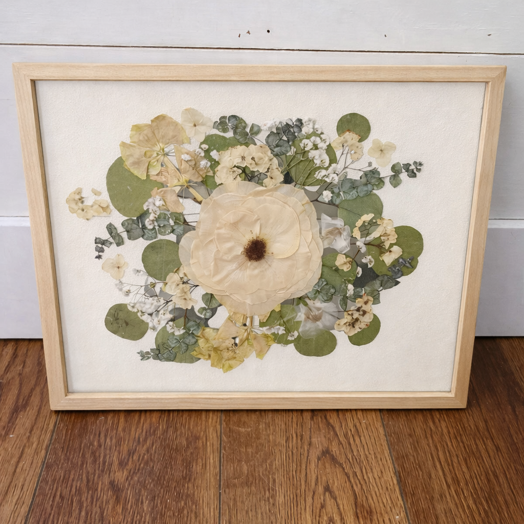 Framed floral arrangement in a natural wood frame with an ivory mat on a wooden floor with a white wall background
