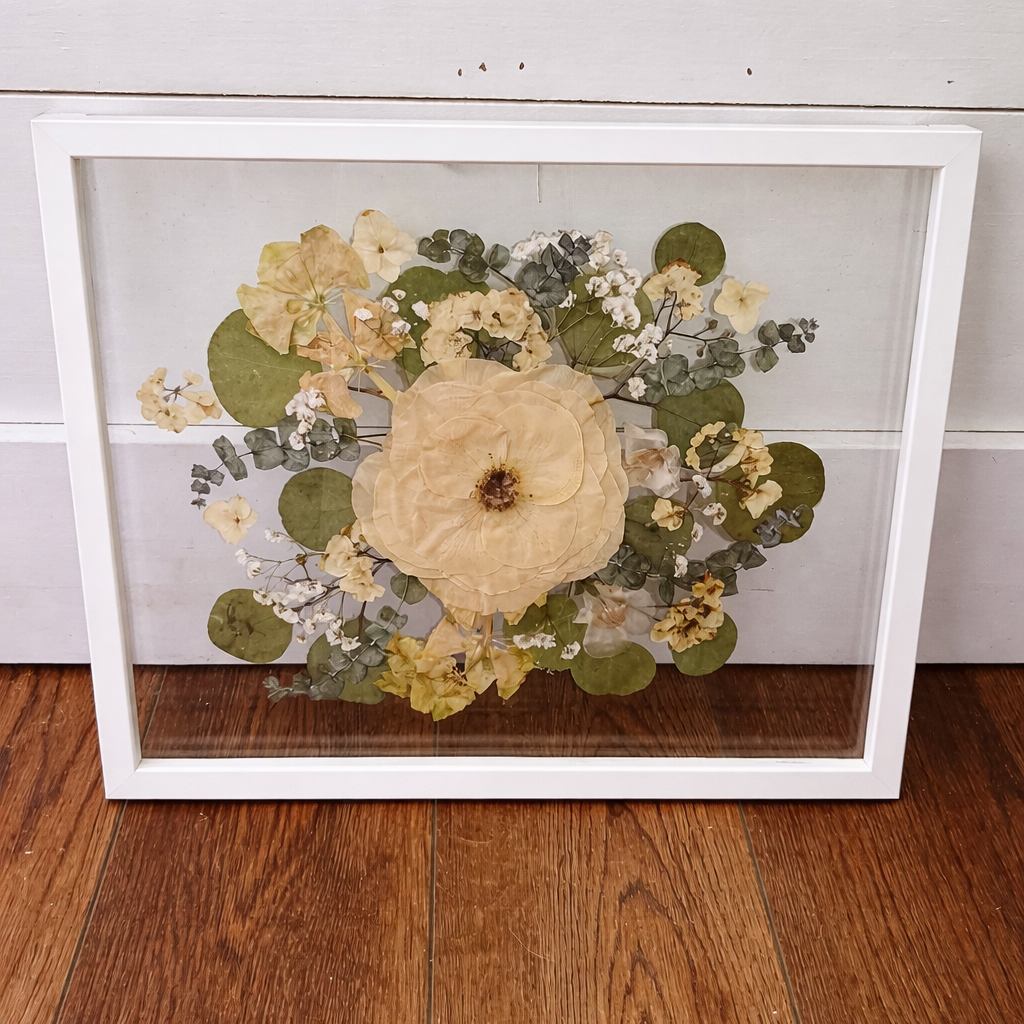 Framed arrangement of pressed flowers in a floating double glass white wooden frame on a wooden floor with a white wall background